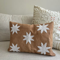 A square tan throw pillow with five white embroidered starburst motifs, resting on a light beige sofa next to another patterned pillow.