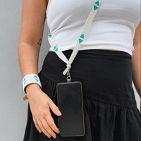 A close-up of a person wearing a white tank top and black skirt, holding a black smartphone in a white fabric pouch attached to a white shoulder strap with teal geometric accents.