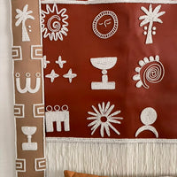 A bohemian-style woven wall hanging tapestry with white Aboriginal-inspired geometric and spiritual symbols on a terracotta red background, suspended from a wooden rod. A matching brown patterned pillow is partially visible at the bottom.