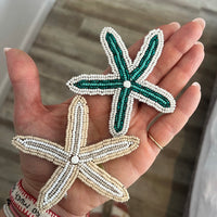 Two hand-beaded starfish charms held in a person's hand. One starfish is green and white, and the other is beige and white.