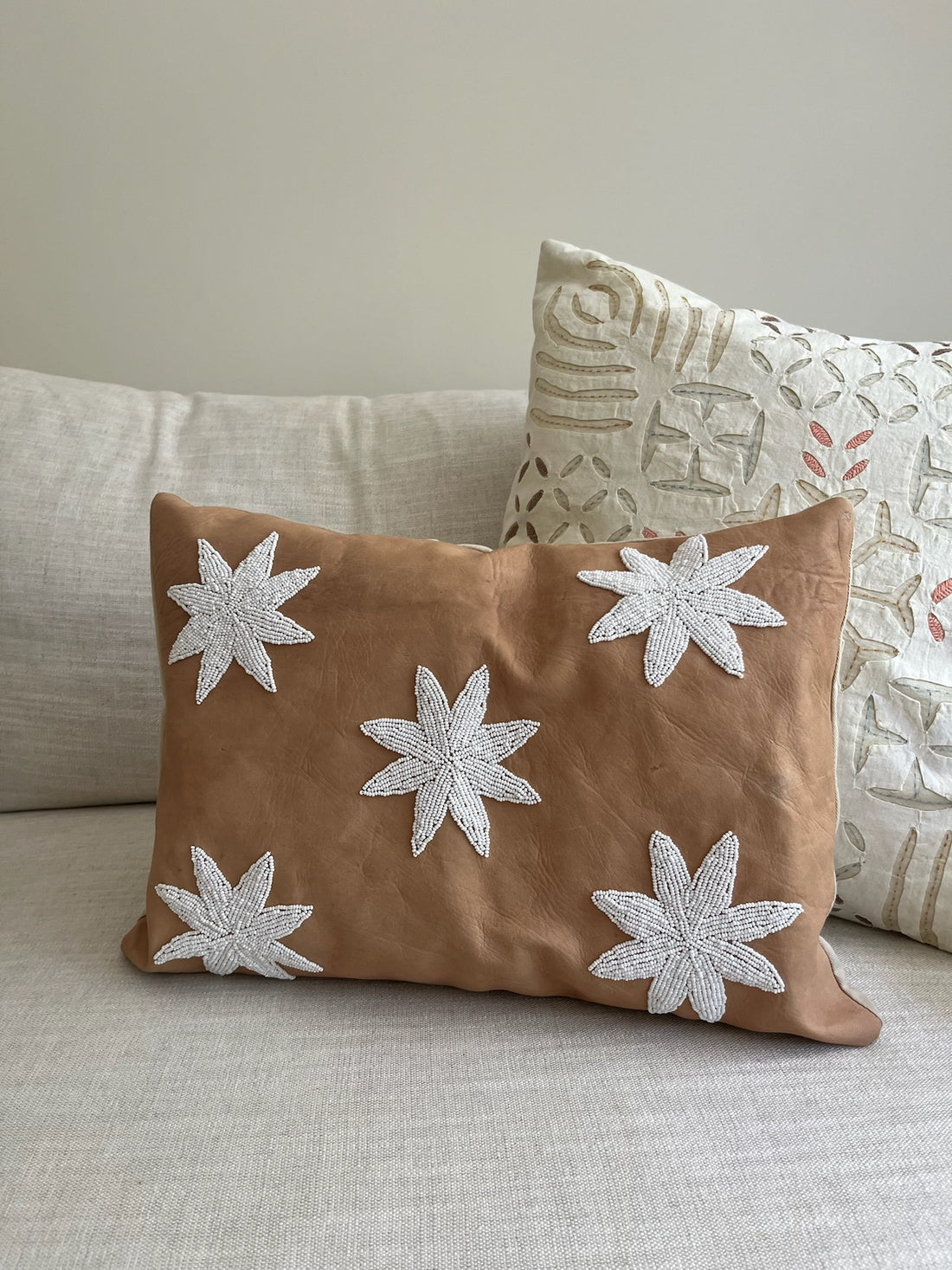 A square tan throw pillow with five white embroidered starburst motifs, resting on a light beige sofa next to another patterned pillow.