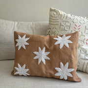 A square tan throw pillow with five white embroidered starburst motifs, resting on a light beige sofa next to another patterned pillow.