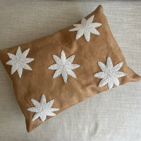 A rectangular tan cushion with five white embroidered starburst motifs, resting on a light beige textured sofa.