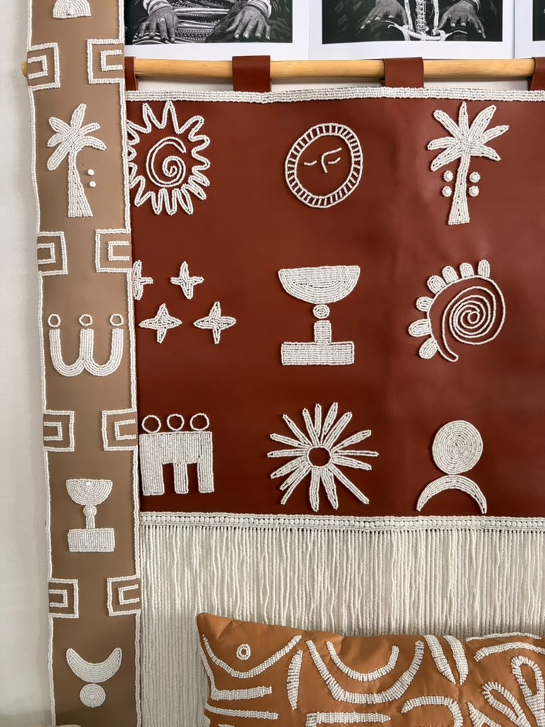 A bohemian-style woven wall hanging tapestry with white Aboriginal-inspired geometric and spiritual symbols on a terracotta red background, suspended from a wooden rod. A matching brown patterned pillow is partially visible at the bottom.