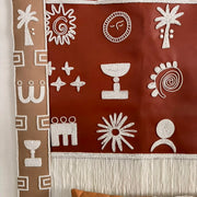 A bohemian-style woven wall hanging tapestry with white Aboriginal-inspired geometric and spiritual symbols on a terracotta red background, suspended from a wooden rod. A matching brown patterned pillow is partially visible at the bottom.