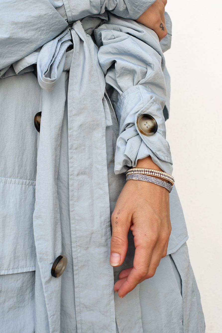 Close-up of a light blue gray coat with buttons and a person's arm wearing a stack of tanzanite bracelets.