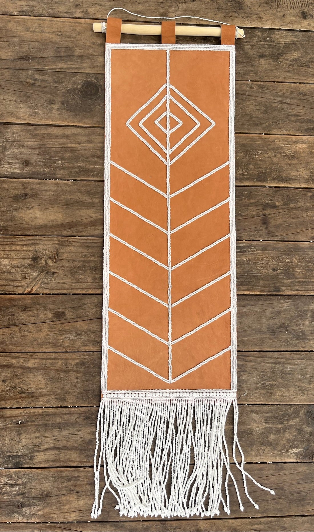 A tan leather and white woven wall hanging with geometric patterns and fringe, suspended from a wooden dowel against a rustic wood plank wall.