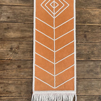 A tan leather and white woven wall hanging with geometric patterns and fringe, suspended from a wooden dowel against a rustic wood plank wall.
