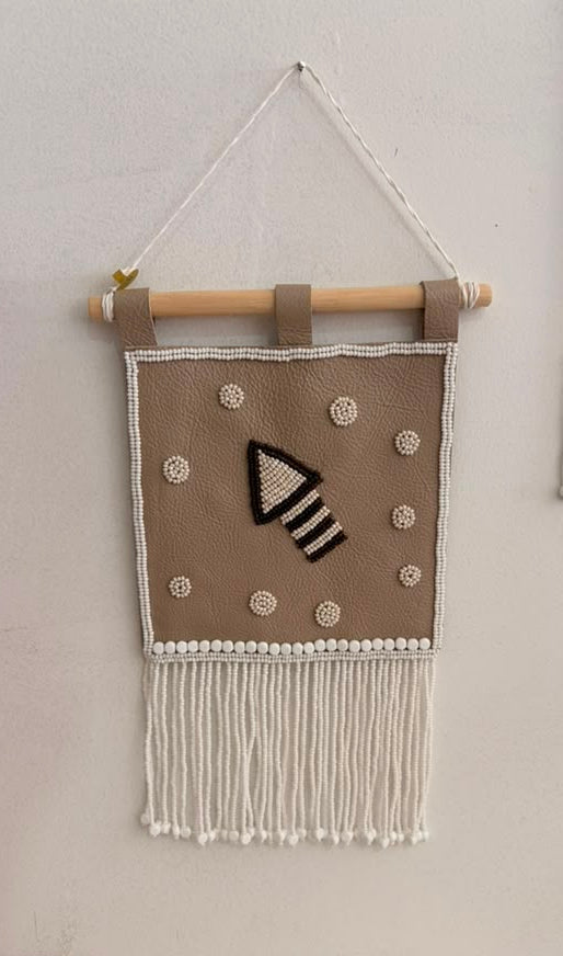A handmade bohemian-style wall hanging tapestry with a beige fabric panel, white fringe, and a black-and-white geometric arrow design, suspended from a wooden dowel against a plain wall.