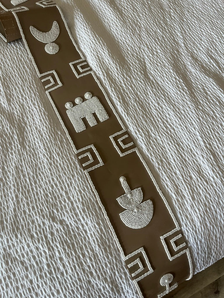 A close-up of a textured cream-colored fabric, likely a quilt or bedspread, featuring a decorative brown vertical band with white geometric and stylized animal motifs.