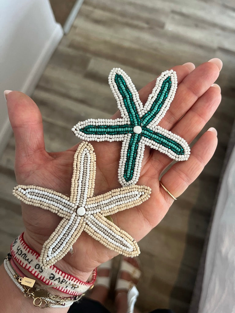 Two hand-beaded starfish charms held in a person's hand. One starfish is green and white, and the other is beige and white.