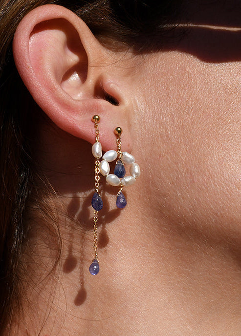 Close-up of a person wearing gold earrings with gemstones and keshi pearls against a neutral background