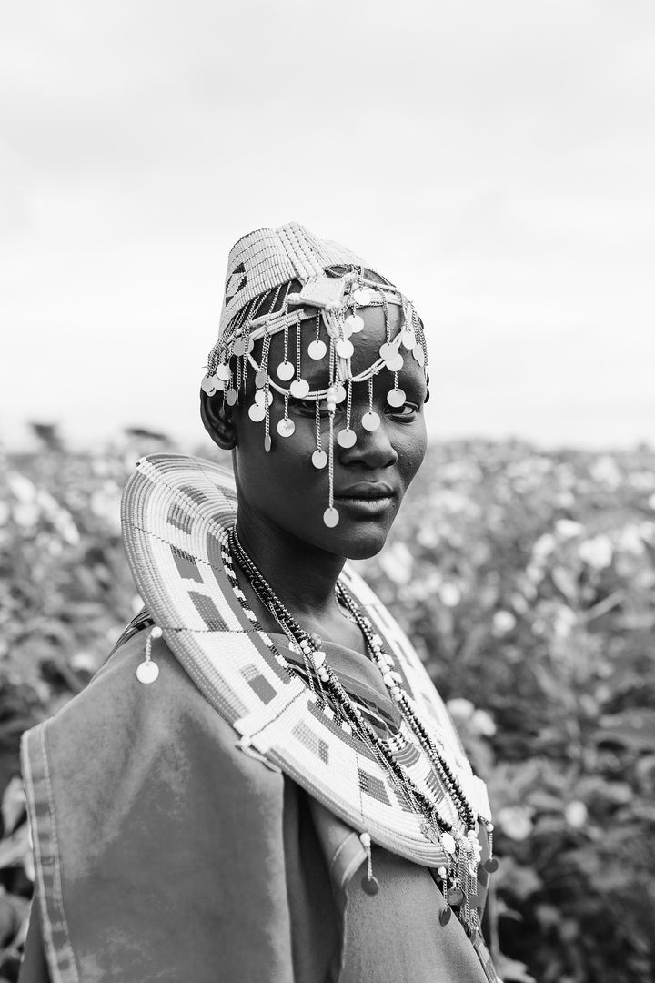 Maasai lady standing in flowering field wearing handmade tribal headpiece, with dangling silver disks and traditional beaded disc.