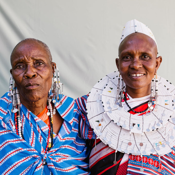 Koko Sadera and Nemasi, Maasai co-wives embracing each other. Dressed in traditional Maasai checked cloth and white beaded disc and headpiece. 