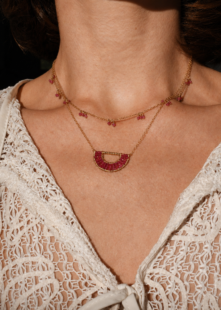 Close-up of a person wearing a delicate gold necklace with red stones against a blurred background.