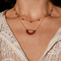 Close-up of a person wearing a delicate gold necklace with red stones against a blurred background.